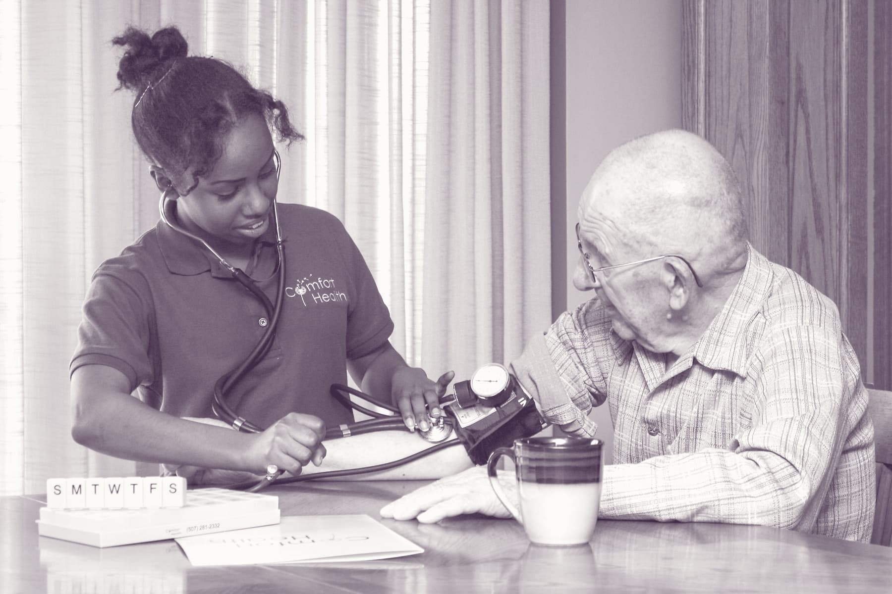A Comfort Health nurse and a longtime Rochester client at his kitchen table — pill organizer and notebook within reach.