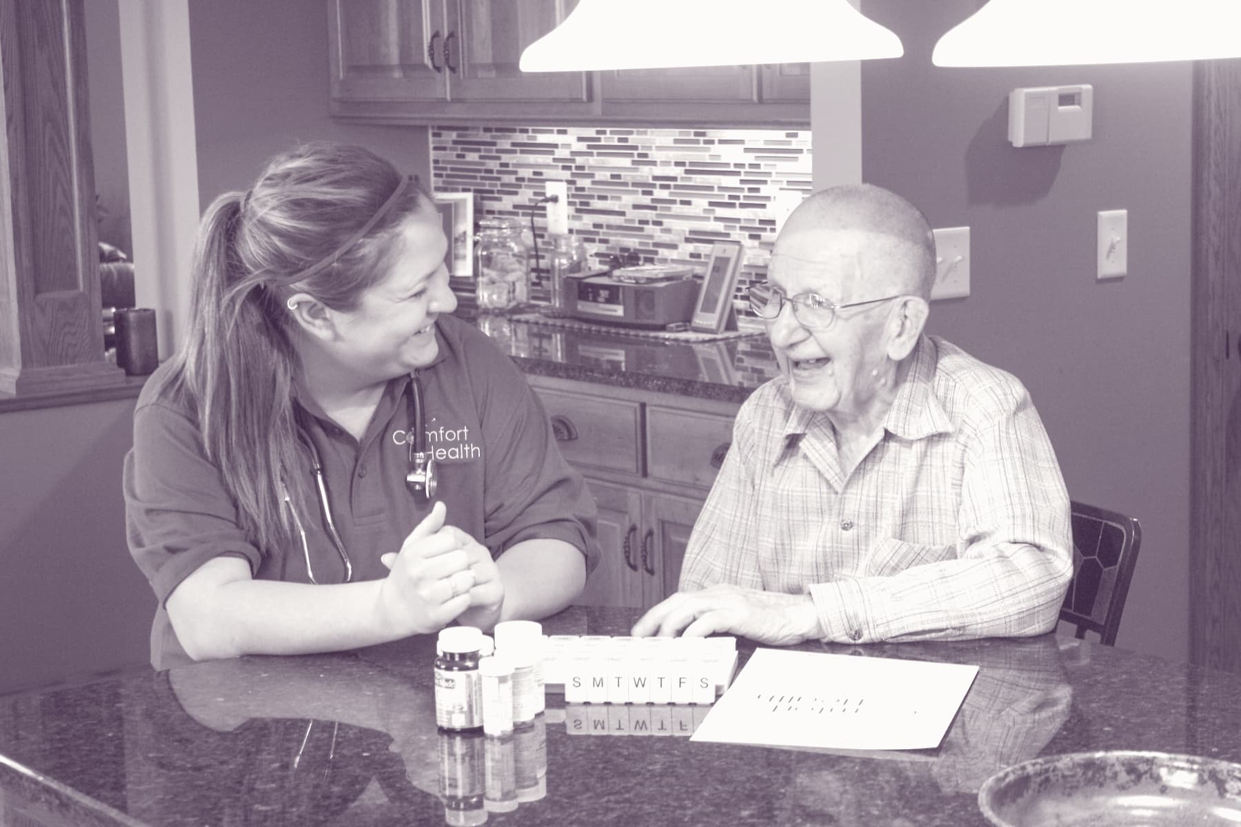 A Comfort Health caregiver laughing with a longtime client at his Rochester kitchen counter, weekly pill organizer between them.