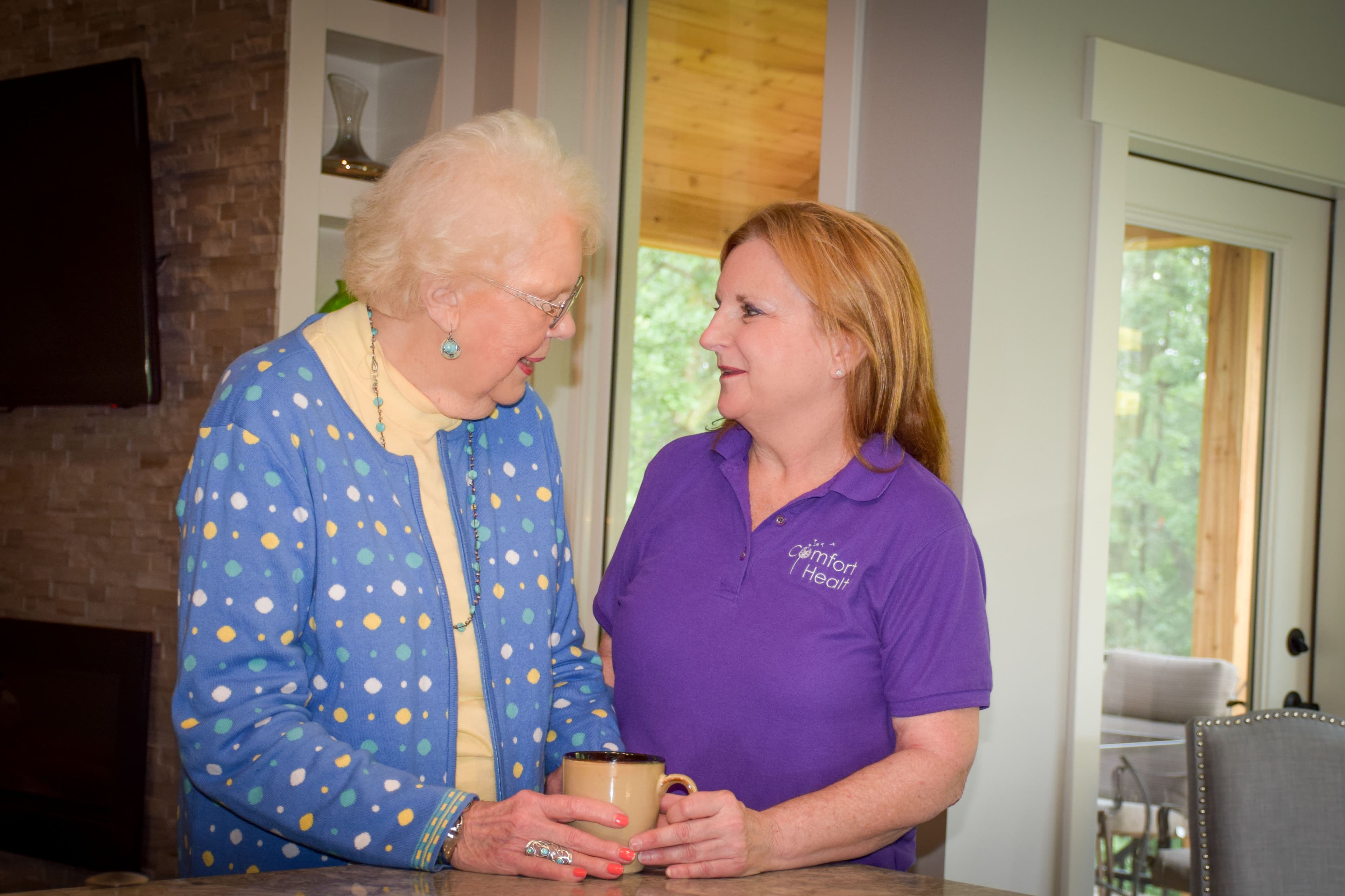 A Comfort Health caregiver in conversation with a Rochester client at her kitchen counter, mid-visit.